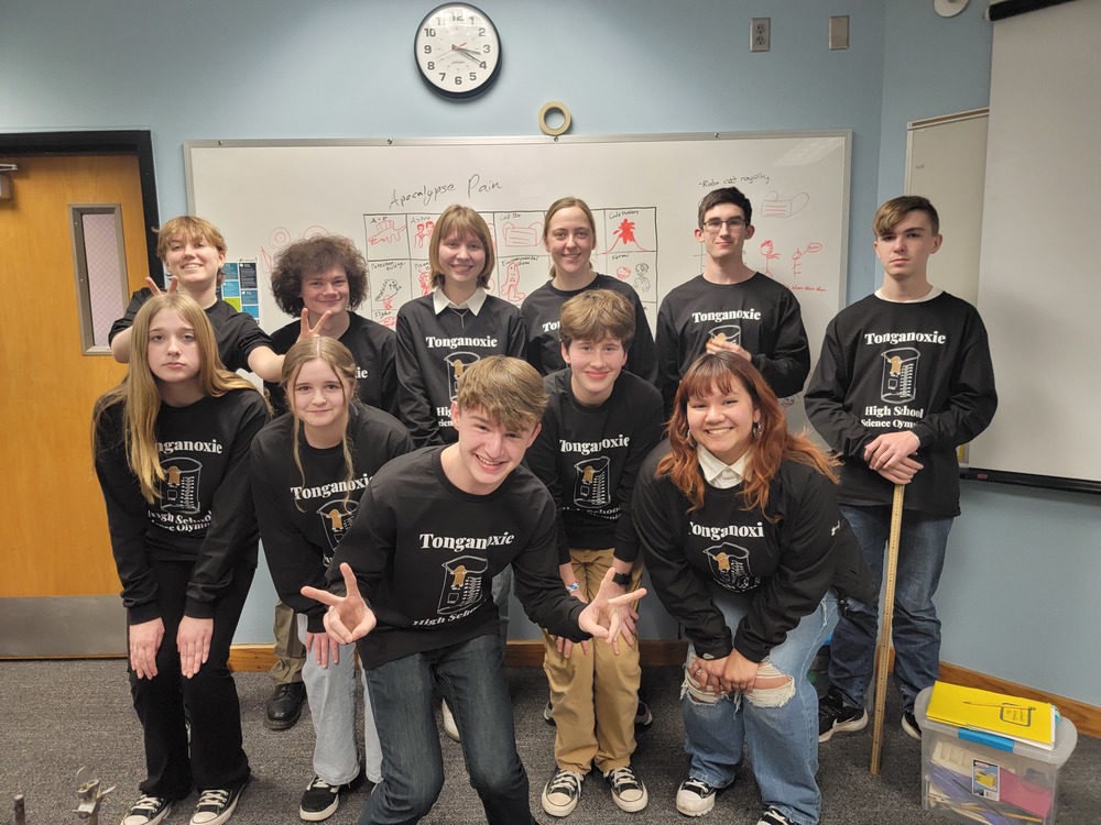 Science Olympiad members pose together in a group in front of a whiteboard, wearing black long sleeved t shirts