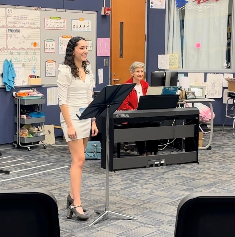 Kyndal Romero wears a white dress and stands behind a music stand. Mrs. Day sits at a keyboard behind her.
