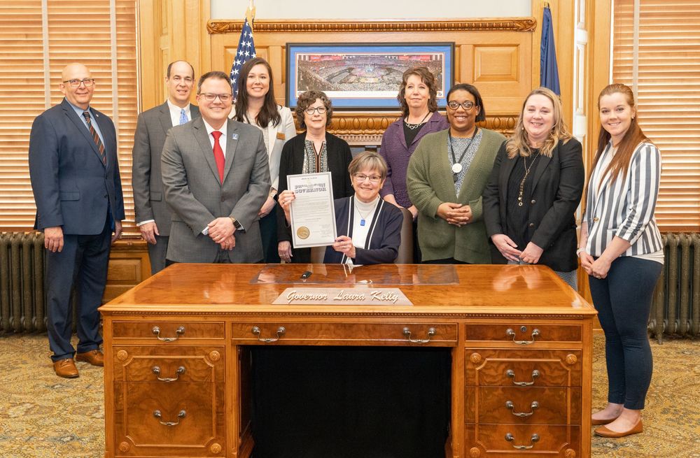 teachers stand behind Governor Kelly as she sits at her desk holding up the proclamation