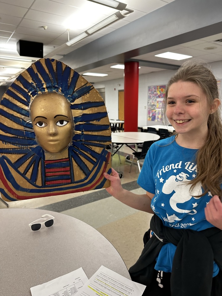 6th grade student wearing a blue t shirt holds a mummy mask she made 