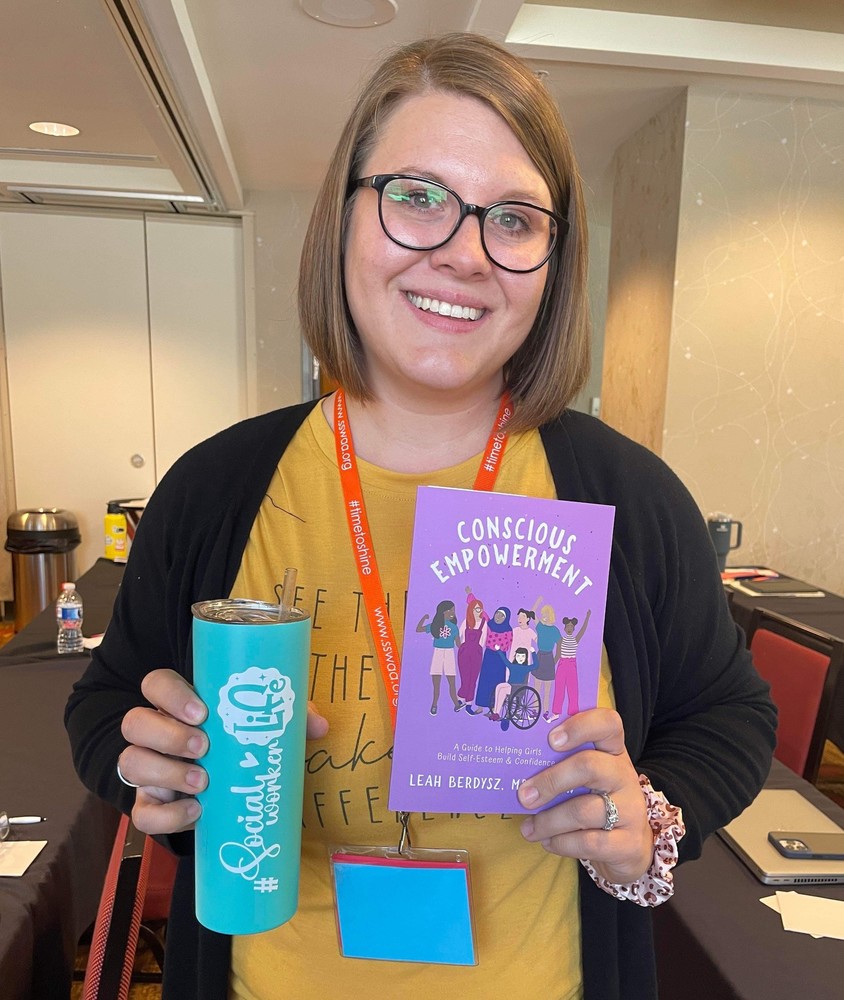 Shelby Burnett wearing a mustard yellow shirt and black cardigan, holding a teal tumbler and purple paperback book