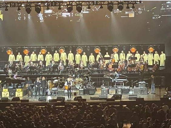 Eagles band members perform on stage. Choir singers wearing yellow robes stand on risers behind them