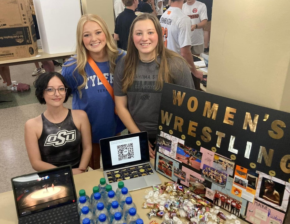 three women standing at a table with water and candy on it, beside  a black poster with gold letters that says women's wrestling