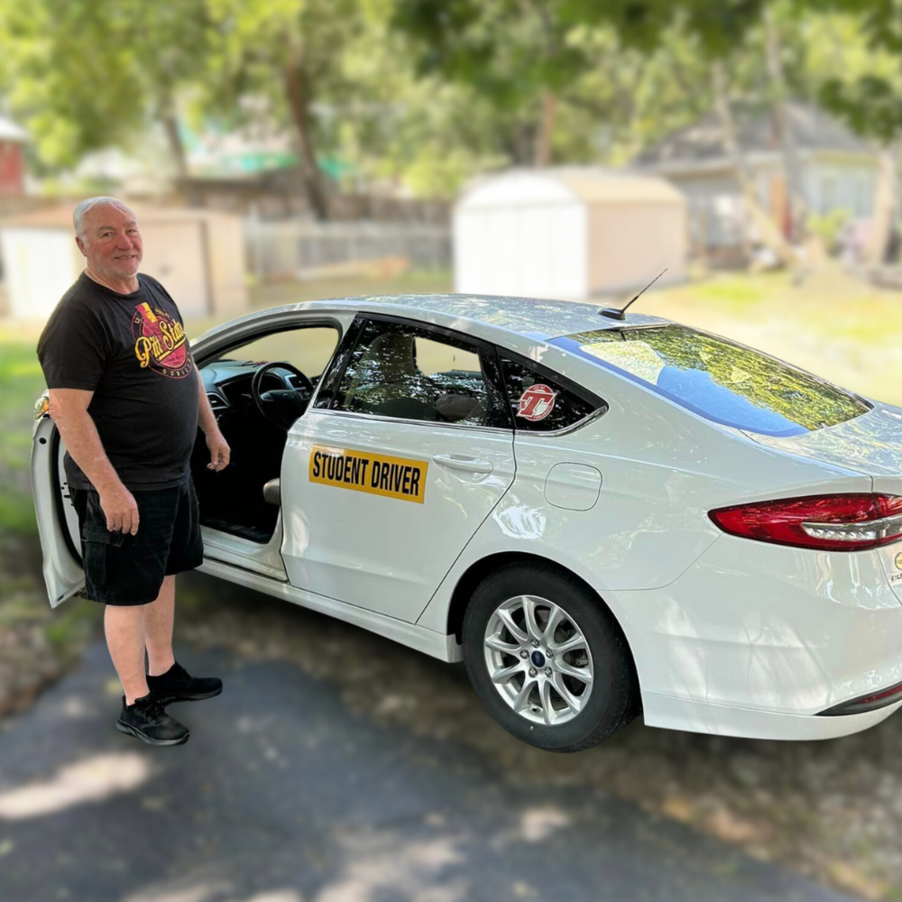 man in black t shirt and shorts stands next to white car with student driver magnet on the back seat driver's side door