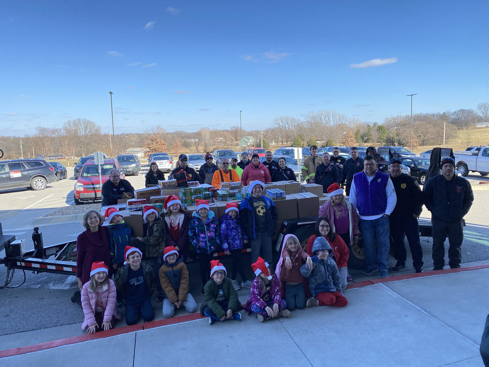 Second grade students sit in front of a trailer filled with donated canned goods