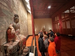 Male students stand on a red tile floor in a museum looking up at a bronze statue