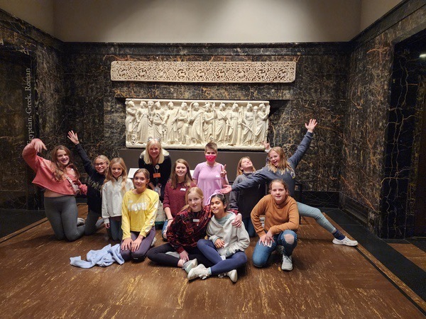 Female students sit on the floor in a museum in front of a carved tablet hanging on the dark brown wall behind them