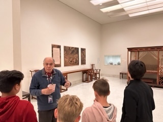 Male students stand in a museum room listening to an older male speak to them