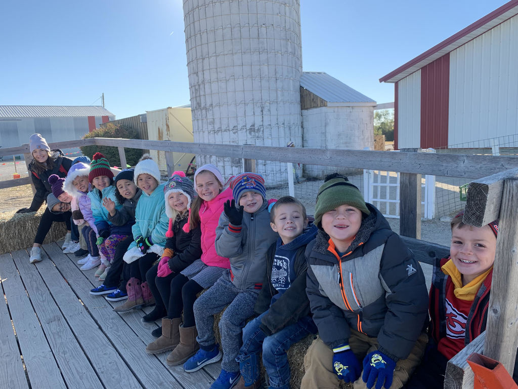 Kindergarten students wearing coats and hats sit on the bench of a hayride truck