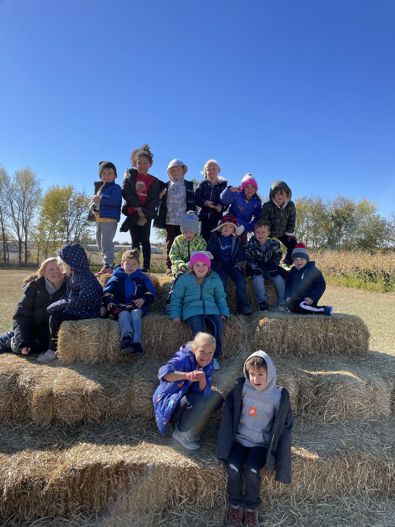 Kindergarten students dressed in coats and hats stand and sit on stacked hay bales