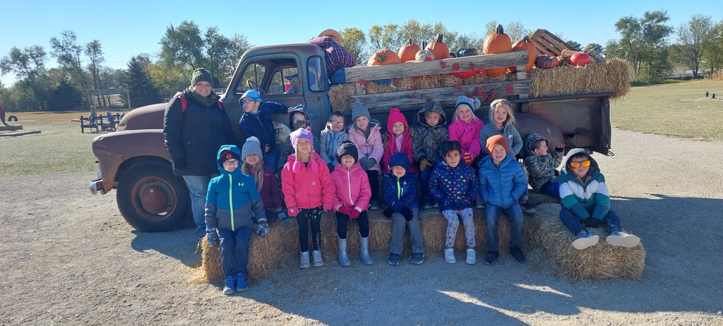 Kindergarten students dressed in coats and hats sit on hay bales stacked in front of  a vintage truck whose bed is full of pumpkins