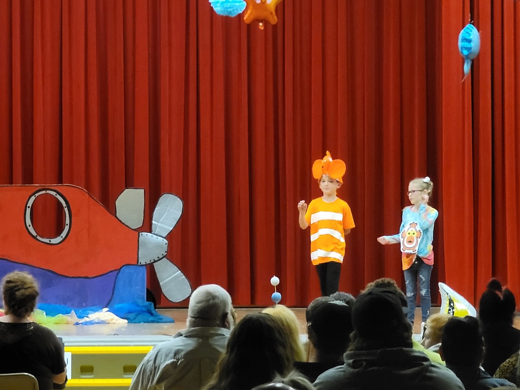 Two second grade students stand on a stage in front of a red curtain; one is dressed like a white and orange clown fish. A cardboard submarine is displayed behind them.
