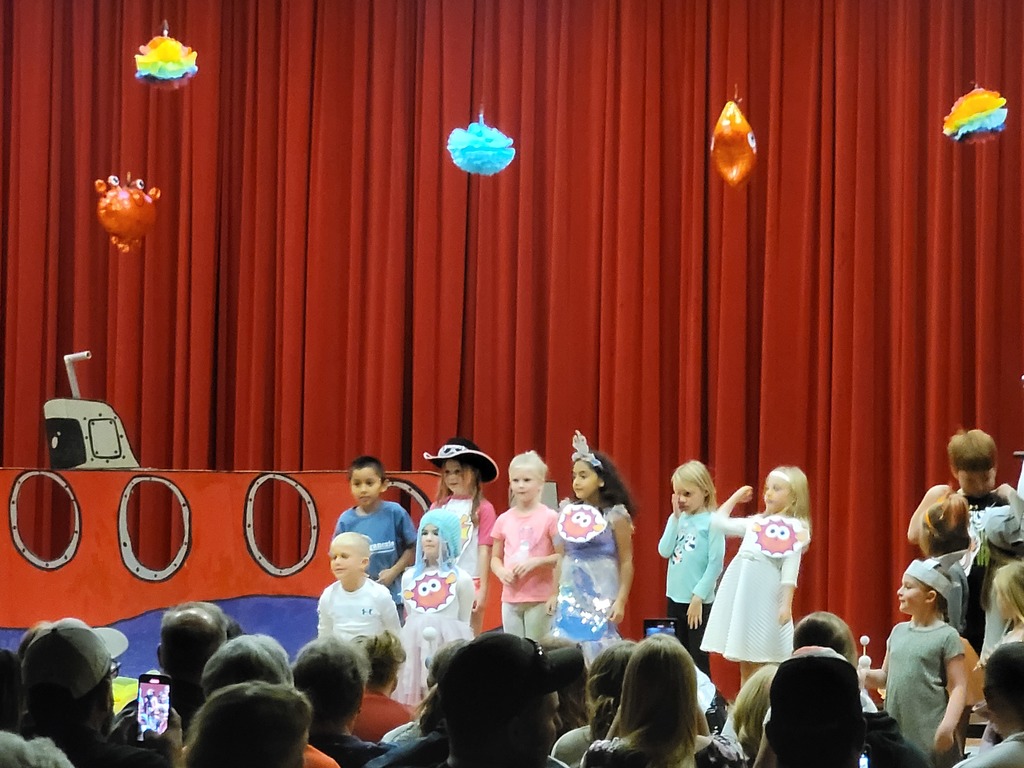 Second grade students stand on a stage in front of a red curtain. A cardboard submarine sits behind them. Several have blowfish signs hanging around their necks.