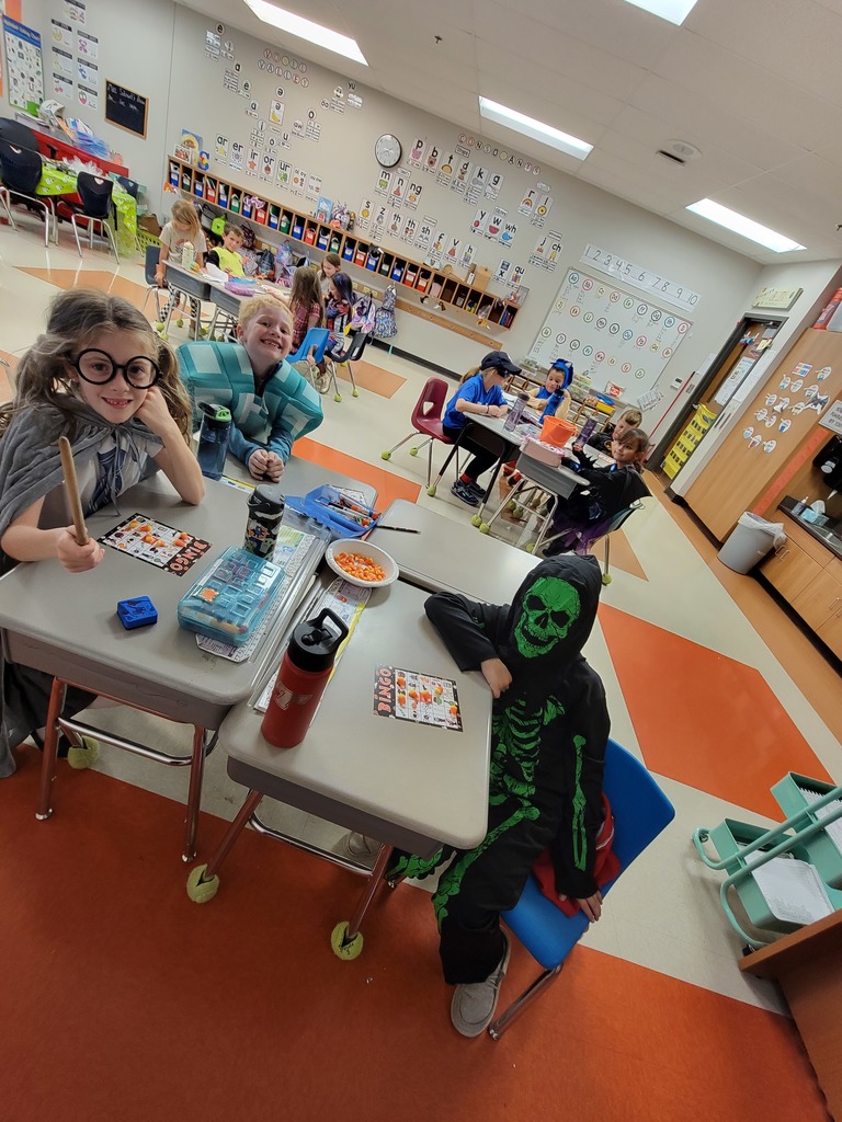 Elementary students sit at their desks in their classroom. Many of them wear Halloween costumes. A Harry Potter character and a black and green skeleton sit in the foreground desks.
