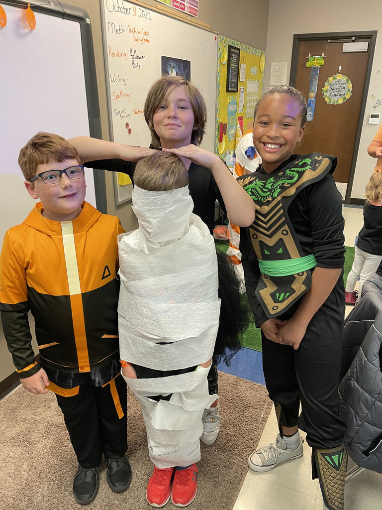 Four elementary students stand together in a classroom. The student in the front is covered head to toe with toilet paper, mimicking a mummy.