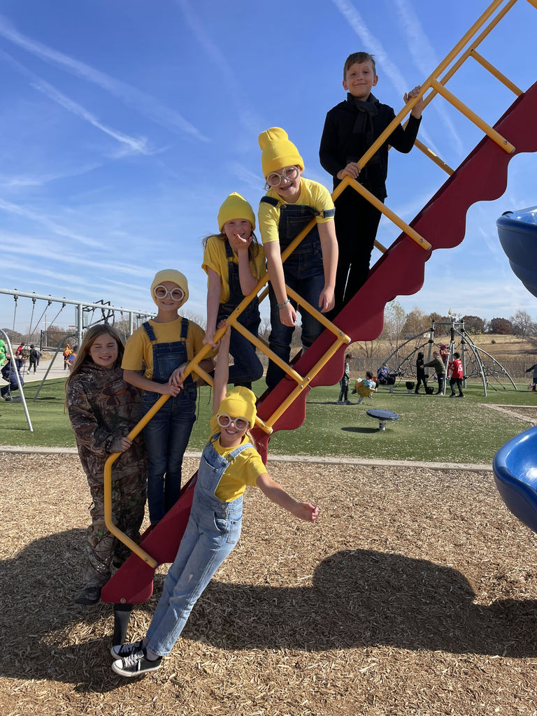 Elementary students stand on the ladder of a playground slide. They are dressed as minions, wearing yellow shirts and stocking caps and denim overalls.