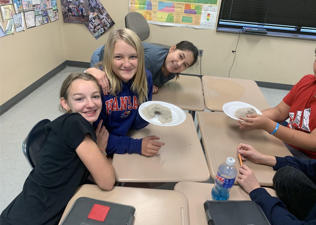 Three seventh grade students sit at desks in a  classroom. One of them wears a blue sweatshirt and holds a styrofoam plate containing kinetic sand.