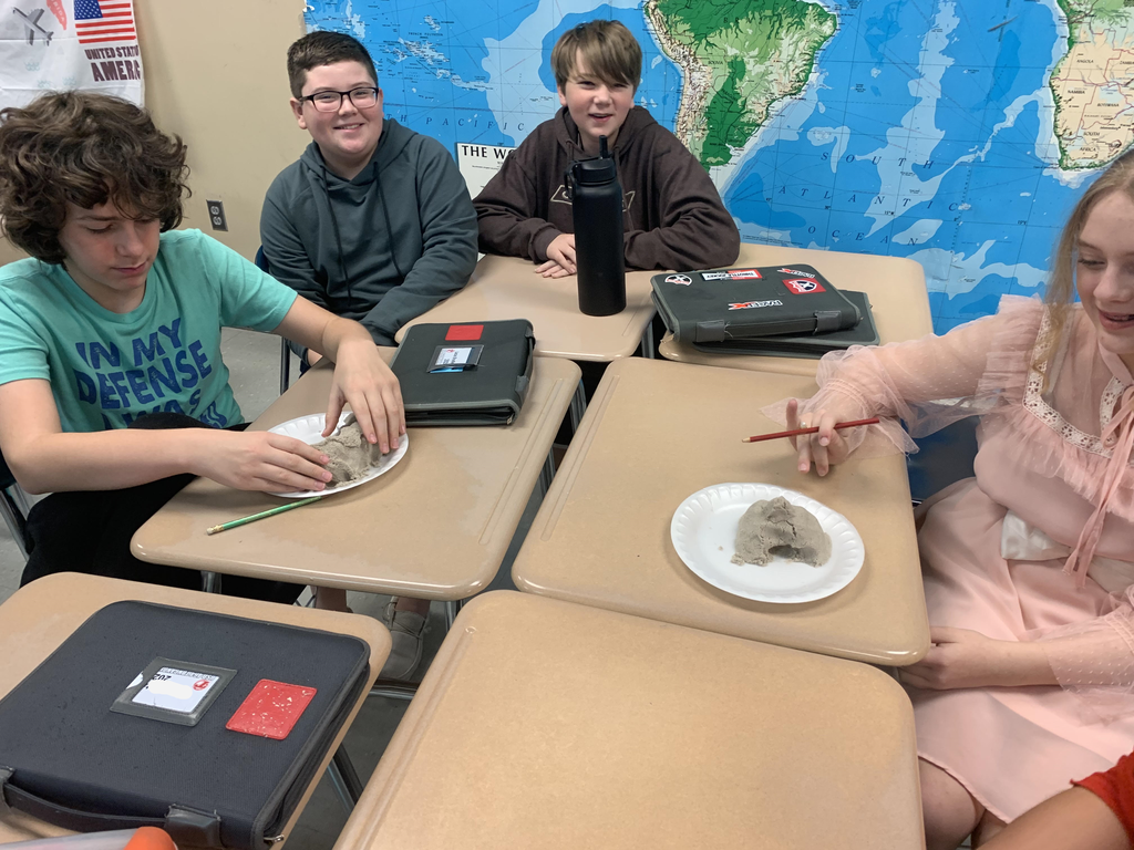 Middle school students sit at desks in a classroom. A world map hangs on the wall behind them. Two students have styrofoam plates containing kinetic sand.