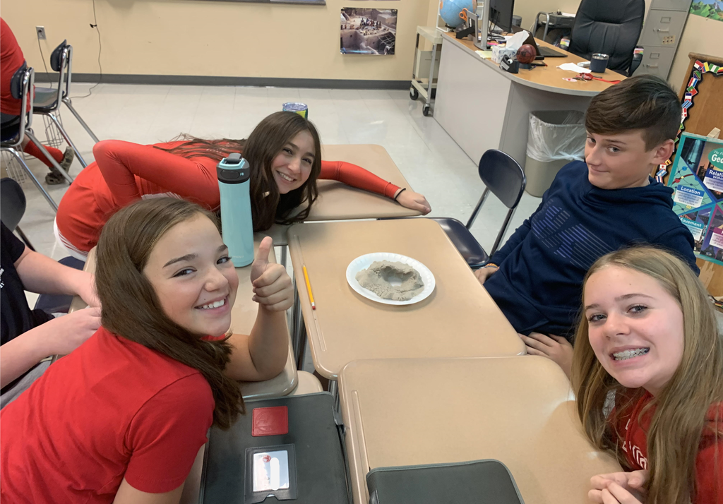 Four middle school students sit at desks in a classroom. One of them has a styrofoam plate containing kinetic sand sitting on the desk in front of him.