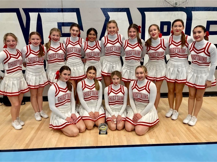 Cheerleaders in white uniforms stand in two rows in a gymnasium. A third place trophy sits on the gym floor in front of them. 