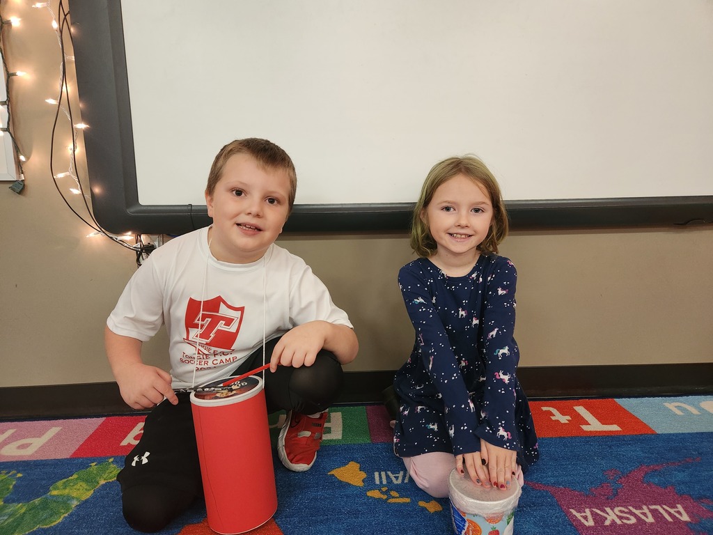 Two first grade students sit on an alphabet carpet in a classroom. Their handmade drums, crafted from oatmeal and food containers, sit on the floor in front of them.
