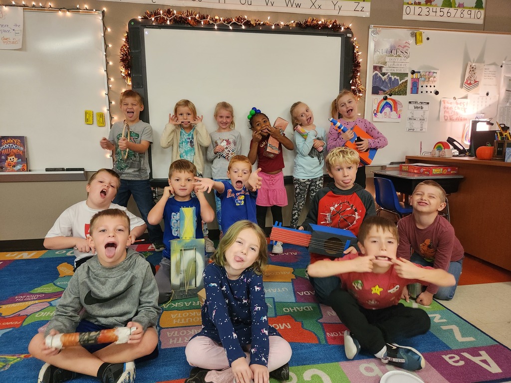 Students sit on an alphabet carpet in a classroom holding their handmade instruments. They are making silly faces.