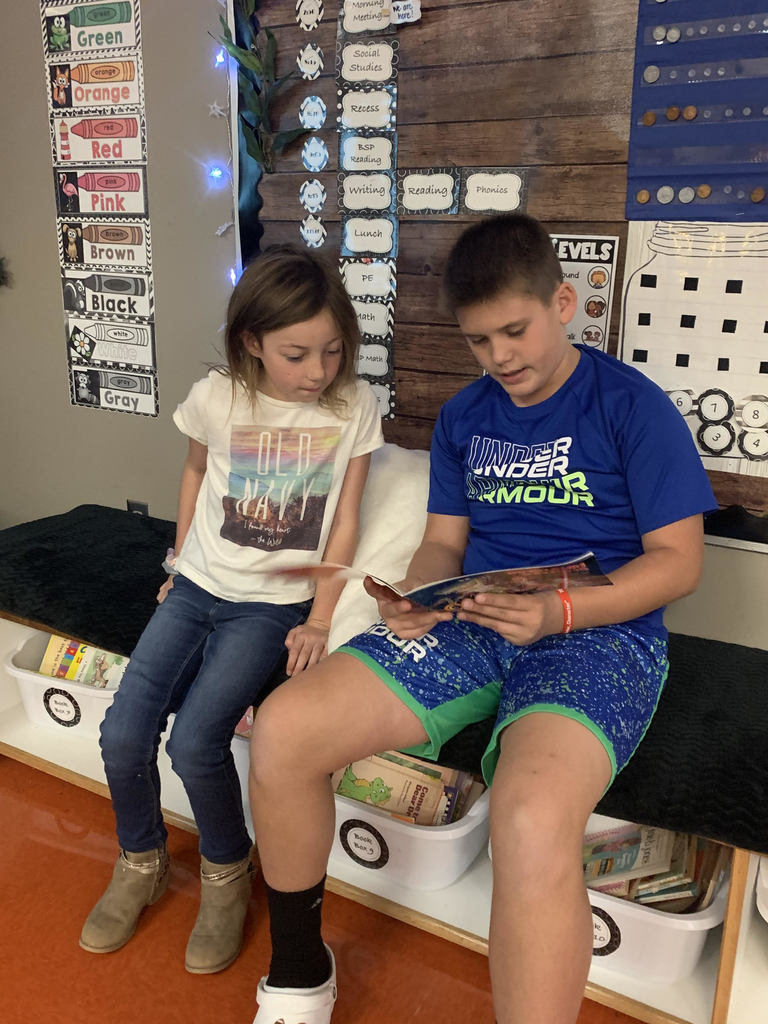 A first grade student and a fifth grade student sit on a bench and read together. The fifth grade student, wearing a blue shirt, holds the paperback book.