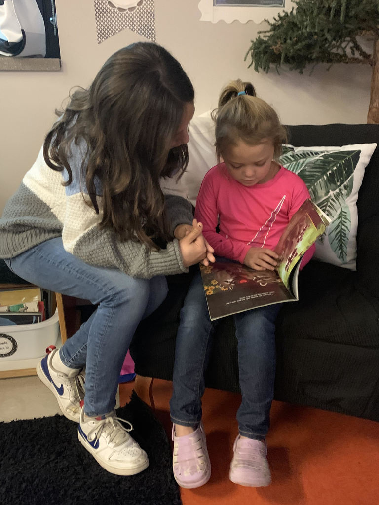 A fifth grade student wearing a green and cream sweater and a first grade student wearing a pink shirt read a book together. The first grade student holds the paperback book.