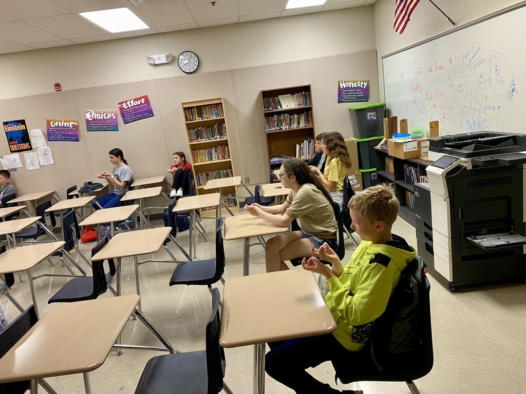 Middle school students sitting in individual desks in a classroom sign various words using ASL.