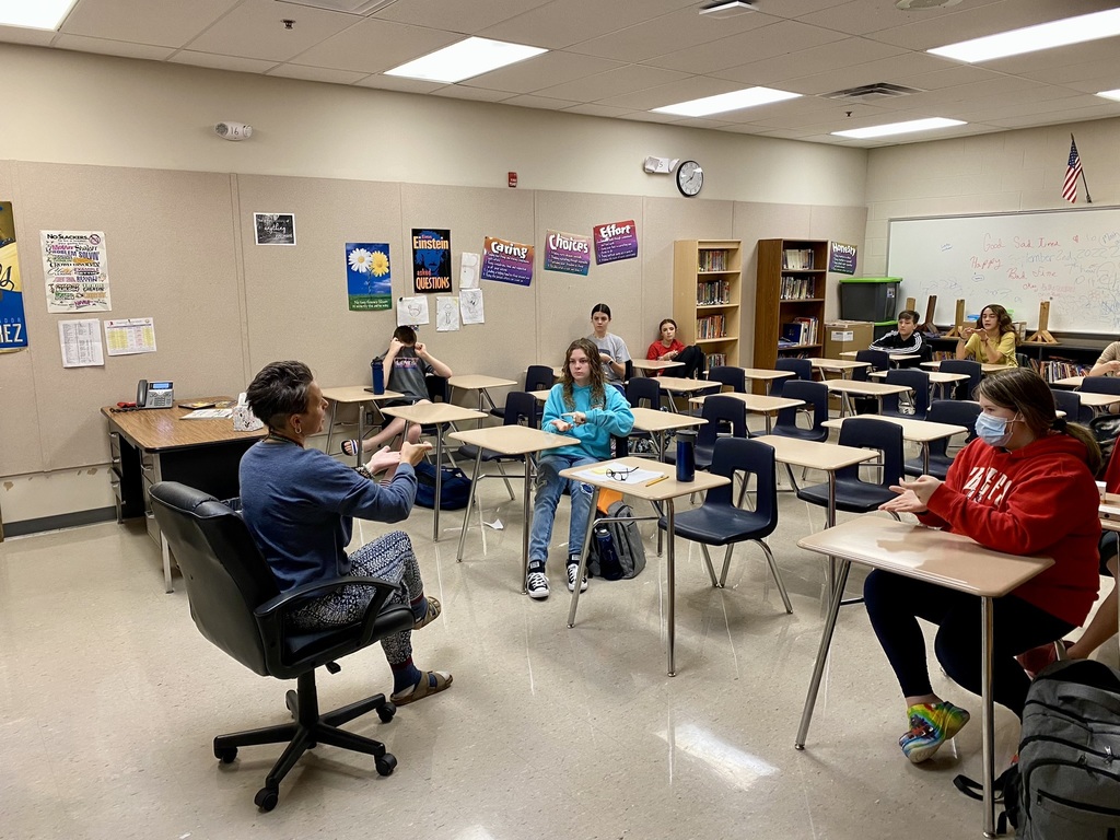 A teacher sits in chair in a classroom and makes a sign with her hands. The students in the classroom, sitting at individual desks, mimic the sign.
