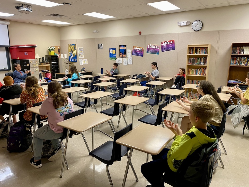 Middle school students sitting in individual desks in a classroom sign various words using ASL.