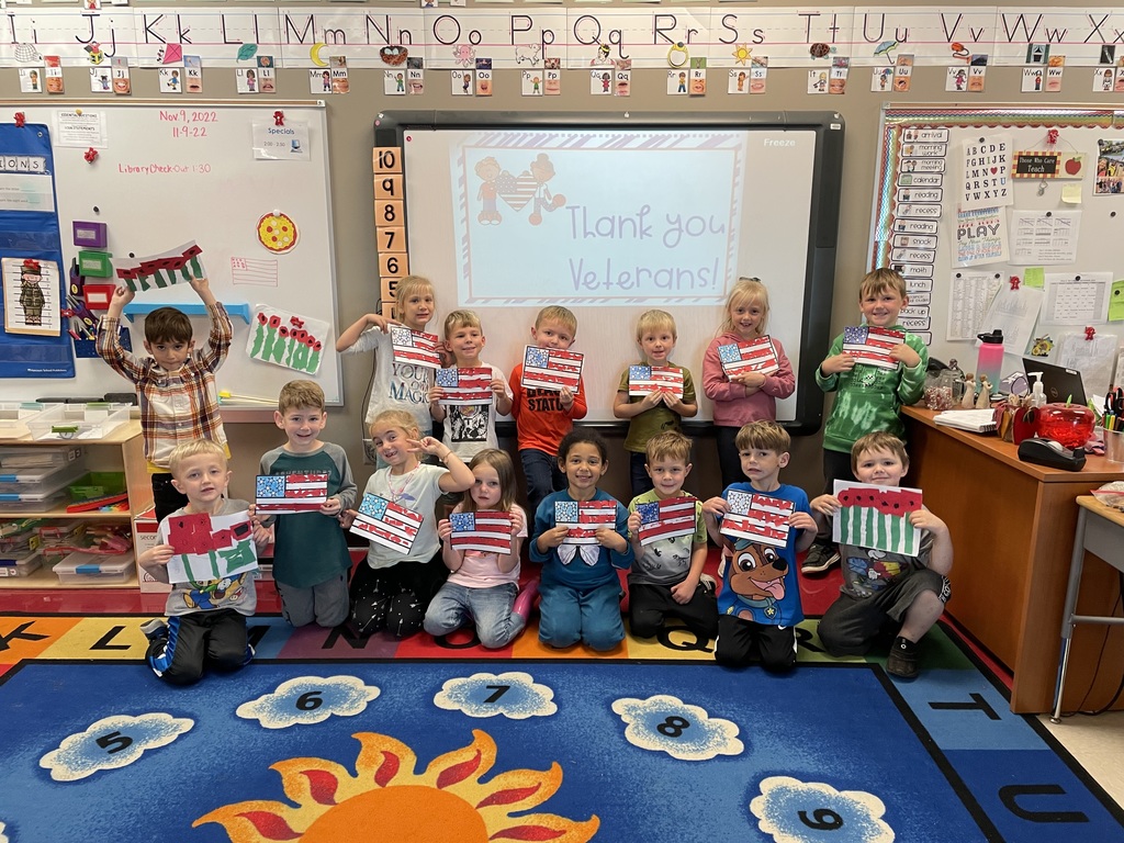 Two rows of kindergarten students stand or sit on a blue rug. They hold American flag themed thank you cards.