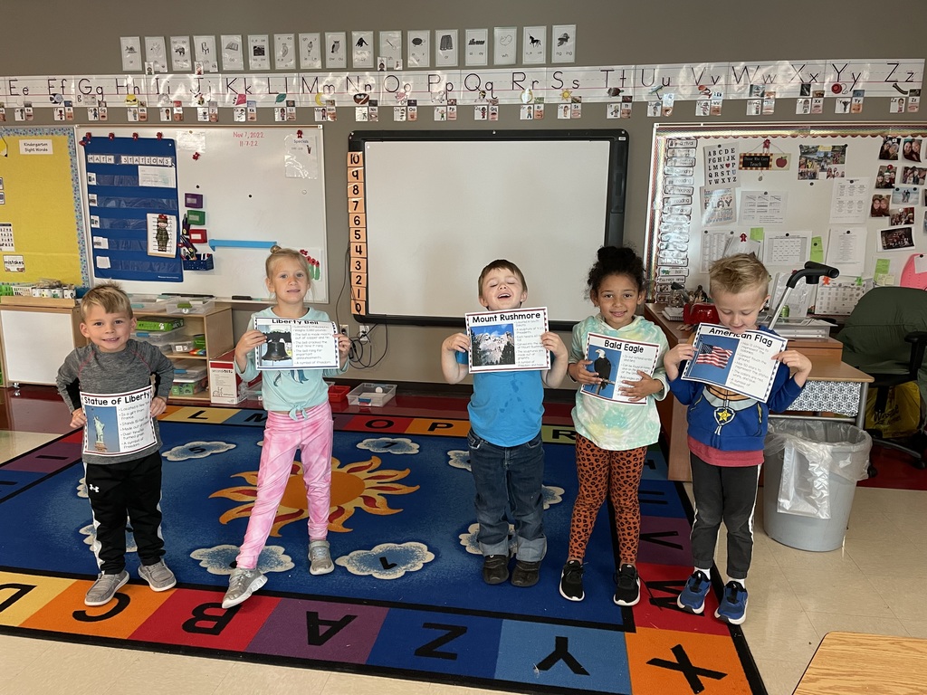 Five kindergarten students stand on a colorful alphabet run in a classroom. Each one holds an 8.5 x 11" flyer depicting a U.S symbol.