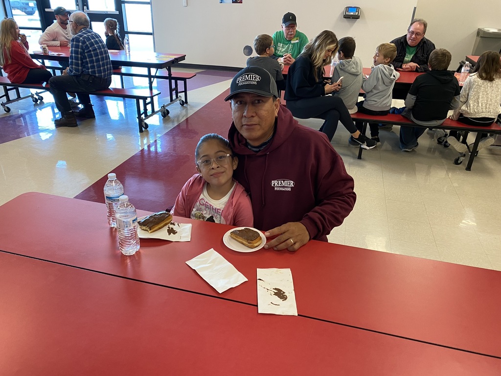 Second grade student and male adult sit together at a red cafeteria table. Donuts sit on the table in front of them.