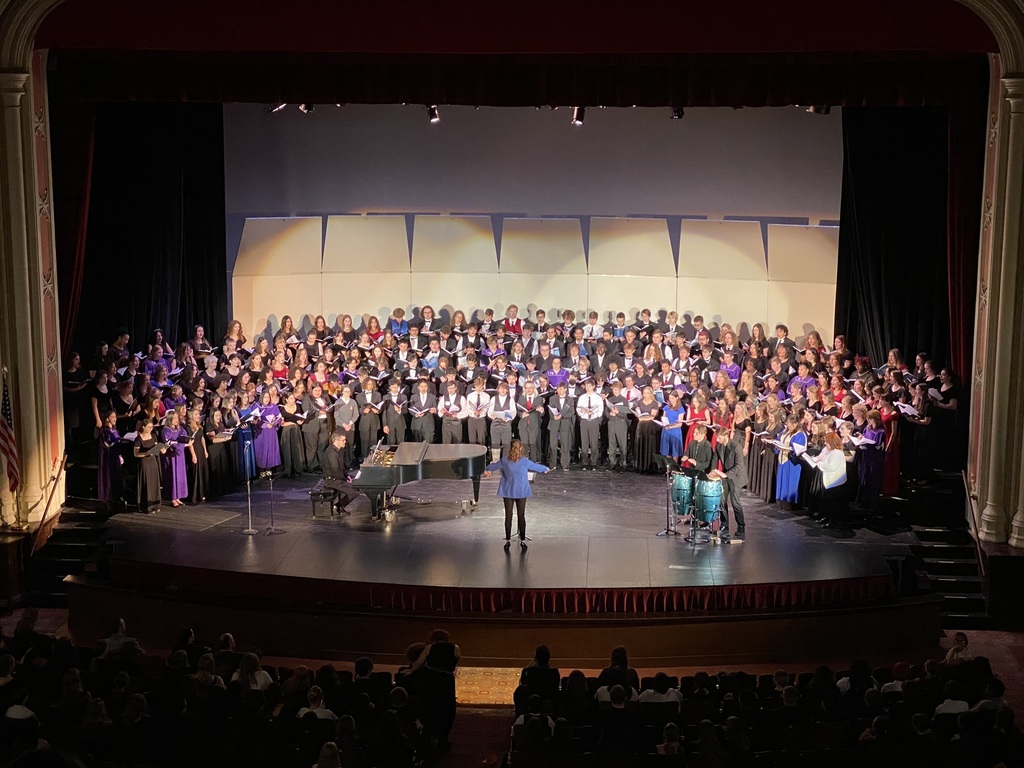 large group of high school singers wearing different colored robes and dresses stand on risers on a stage while a woman in blue blazer conducts