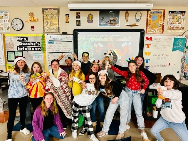 group of students wearing santa hats, reindeer antlers, standing in a classroom in front of a smart board