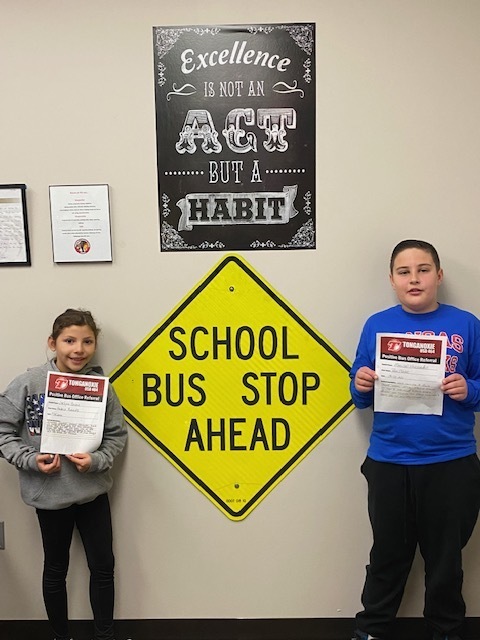 two elementary students stand in front of a wall holding positive bus referral certificates. a traffic sign on the wall says "school bus stop ahead"