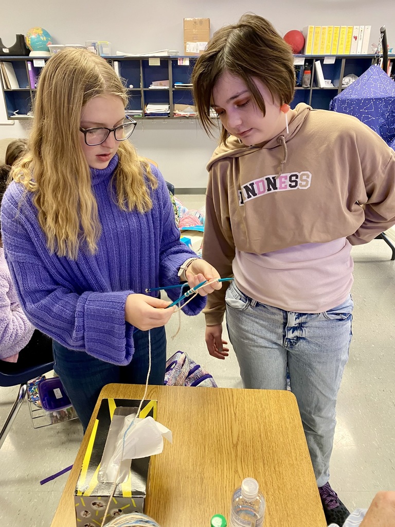 a student wearing a purple sweater shows  a student wearing a brown hoodie how to knit