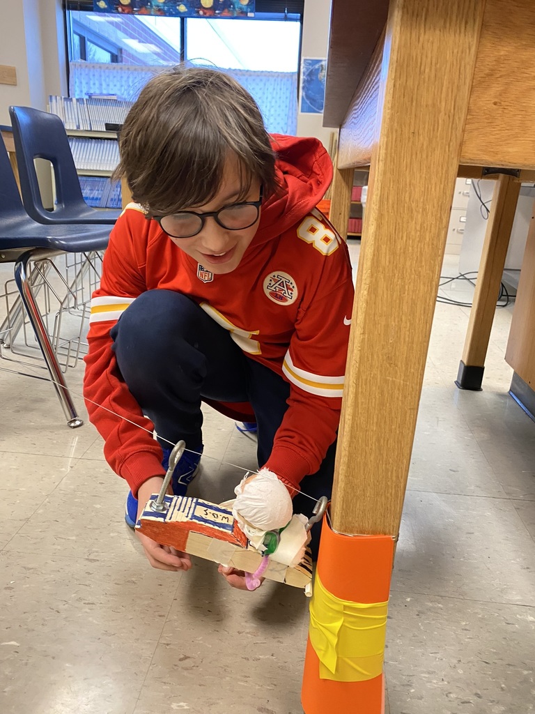 a student with brown hair wearing a Chiefs jersey crouches down to collect his pinewood derby plane after it crashes into a table leg