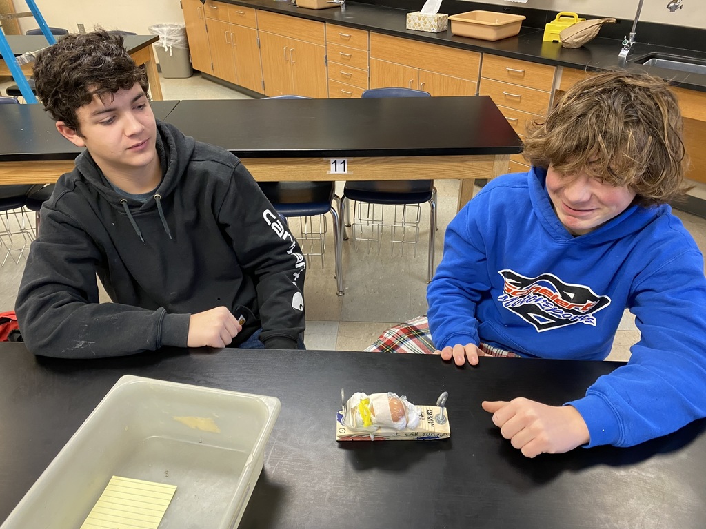 two middle school students sit at a table. their pinewood derby plane with egg strapped to it sits on the table in front of them