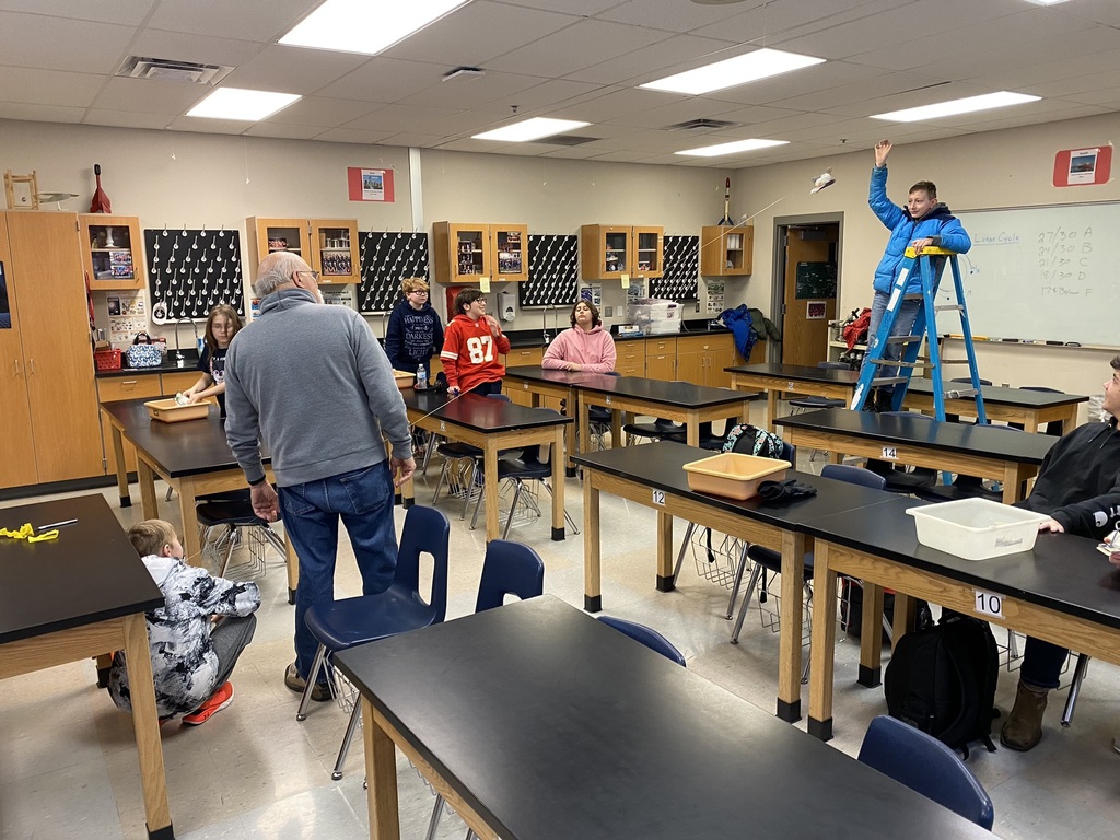 a student wearing a blue coat and standing on a ladder lets go of his pinewood derby plane that is suspended from the ceiling with fishing wire