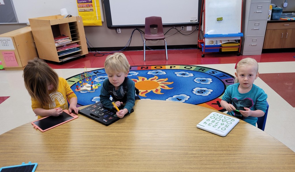 students sit at a brown half circle table using tracing and writing tools to practice their letters, numbers, and shapes