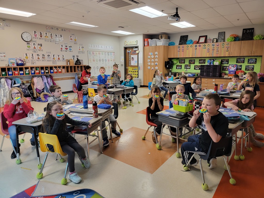 first grade students sit at their desks in their classroom eating sugar cookies frosted with blue icing and yellow sunflowers