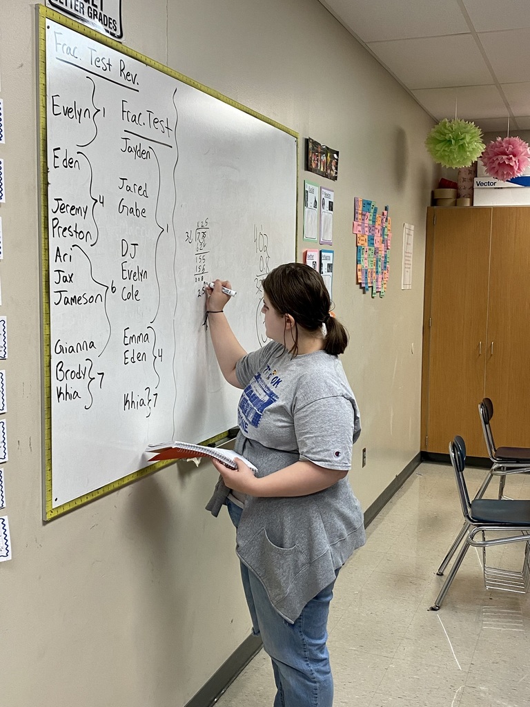 student wearing a gray t shirt stands at a white board working on a fraction division problem