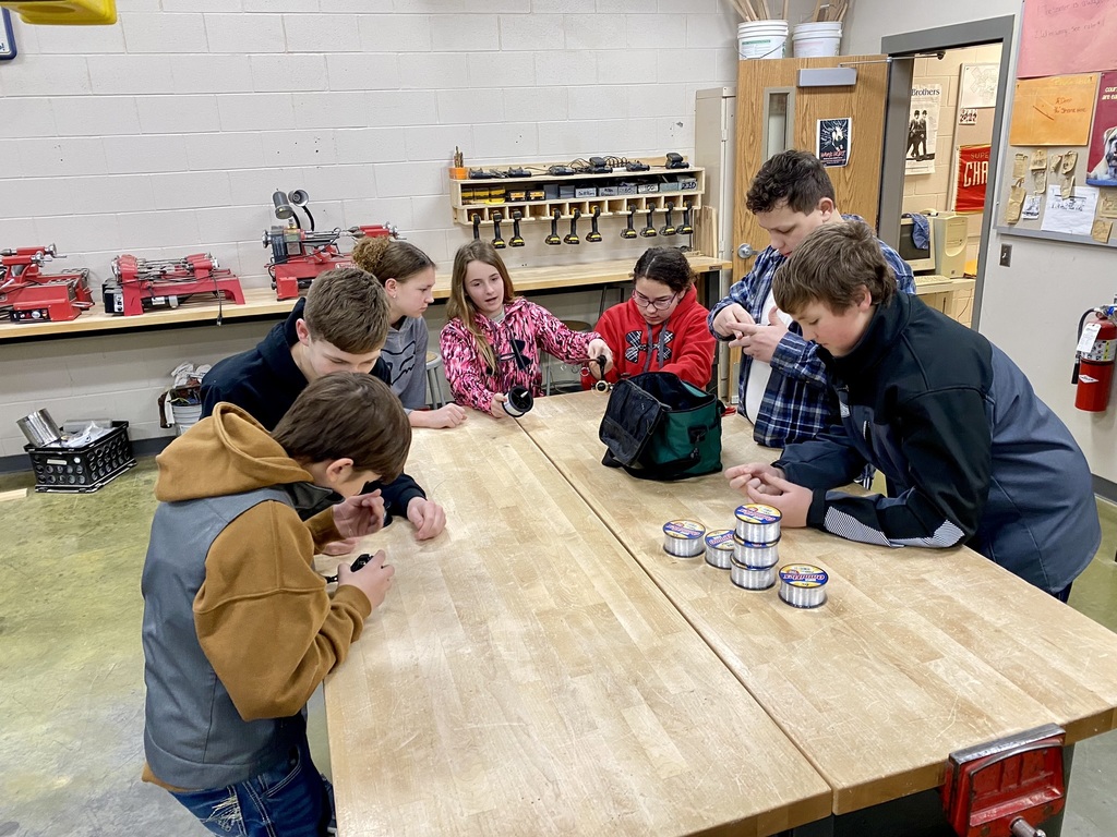 a group of middle school students stand around a wooden topped table and work on putting fishing line on a reel
