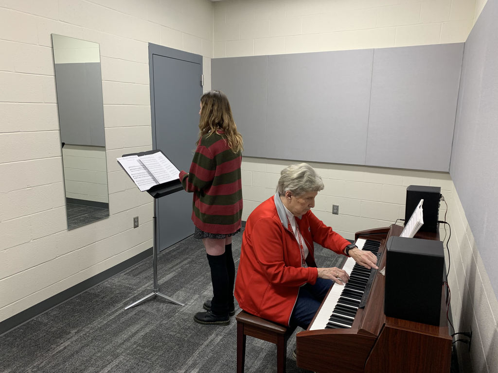 Josey stands before a wall mirror with a music stand in front of her. Mrs. Day wears a red sweater and sits at a keyboard.