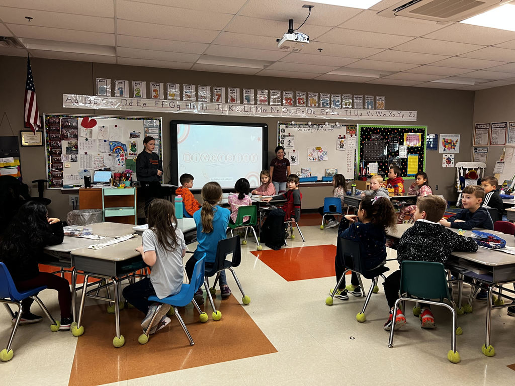two middle school students stand in front of a classroom by a smartboard. first graders listen as they speak about diversity