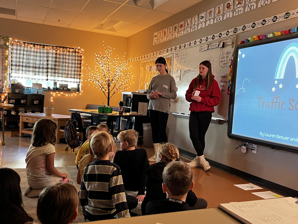 two middle school students stand in front of a classroom next to a smartboard that says traffic safety. second graders sit on the carpet in front of them
