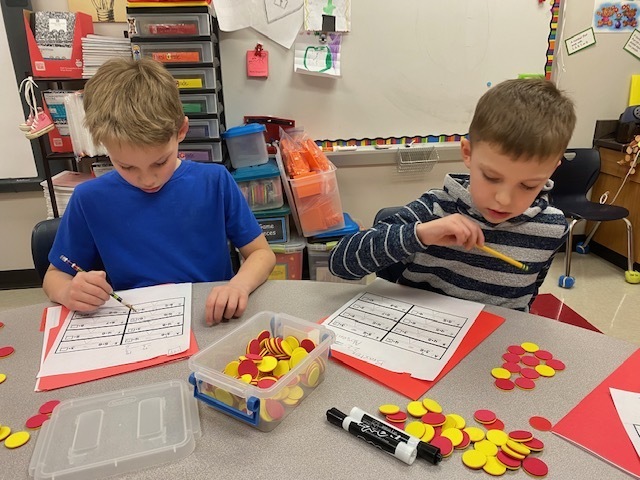 two students work on a math worksheet. yellow and red counters sit on the table.