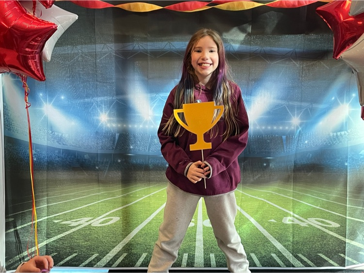 fourth grader holds paper gold trophy standing in front of football field trophy  